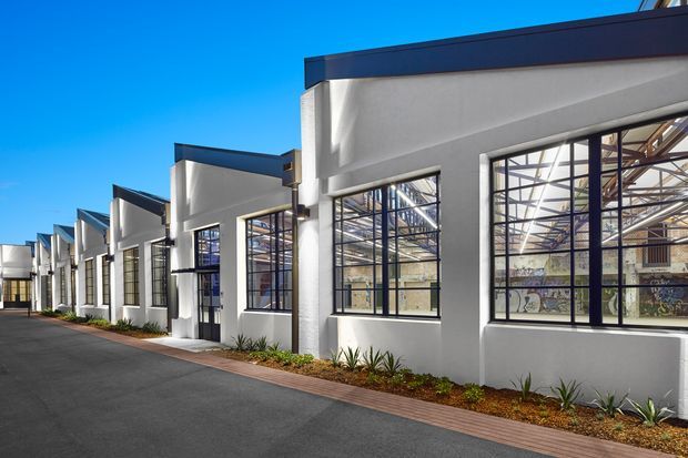 Exterior view of a contemporary low-rise commercial or industrial building with a repeating sawtooth-style roofline, white rendered walls, dark metal-framed windows, and exposed structural elements visible through the glazing. A paved driveway and narrow landscaped planting strip run alongside the facade under a clear blue sky.