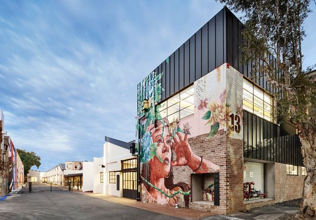 Exterior view of a contemporary multi-storey commercial or creative-industrial building in an urban setting. The building combines dark vertical metal cladding, exposed brickwork, expansive glazed openings, and a large colourful mural wrapping the facade. A gated paved access lane and adjoining low-rise buildings are visible, with mature trees beside the structure.