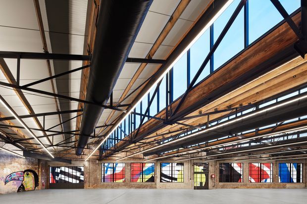 Large open-plan industrial interior with exposed timber roof framing, black steel trusses, extensive exposed ductwork, concrete flooring, brick walls, and tall steel-framed clerestory glazing. Colourful graphic panels are visible along the lower perimeter walls, with strong daylight entering from above.