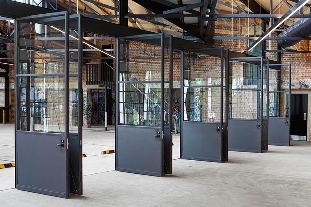 A spacious adaptive-reuse industrial interior with rows of dark metal-framed glazed partitions and doors. Exposed red-brick walls, exposed steel roof structure, visible services, and a polished concrete floor create a warehouse or studio-like commercial workspace. The open-plan layout is divided into enclosed rooms with extensive internal glazing.