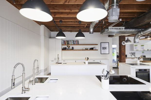 A bright contemporary commercial-style kitchen or studio workspace with extensive white island benches, multiple sinks and taps, induction cooktops, white cabinetry, open shelving, and stainless-steel appliances. The space has an exposed timber beam ceiling with exposed metal ductwork and large black pendant lights, creating a clean industrial aesthetic.