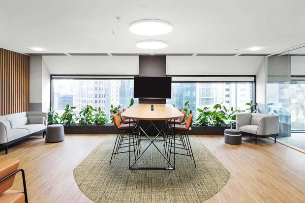 Bright contemporary office lounge and meeting area with a round timber meeting table, black wire-frame chairs, sofas, armchairs, indoor plants, and a wall-mounted screen. The space has pale timber flooring, a large neutral woven rug, full-height city-facing windows, timber slat wall detailing, and glass partitions.