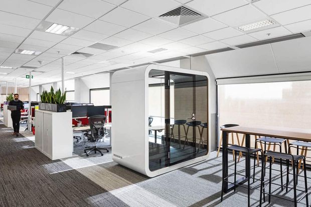 Bright contemporary open-plan office interior with workstation clusters, a freestanding enclosed meeting pod with curved white walls and glazed doors, and a high communal bar table with stools beside large windows. The space has a suspended acoustic tile ceiling, carpeted flooring, and a clean corporate fit-out.