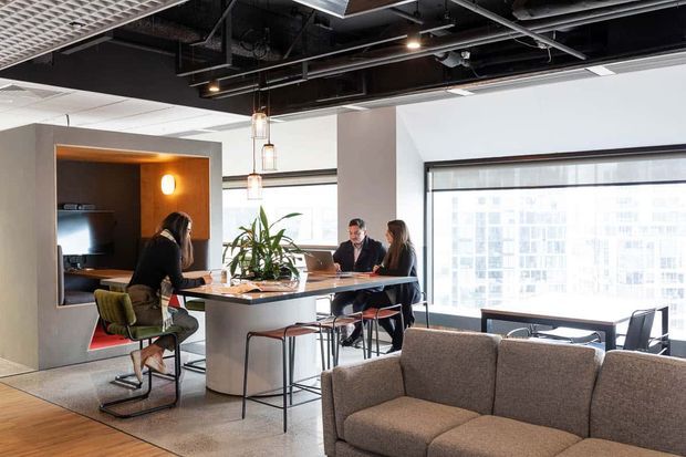 A bright contemporary open-plan commercial interior with a central communal island and high stools, a small kitchenette or meeting nook, exposed black ceiling services, pendant lights, large windows, and a partially visible upholstered sofa seating area.