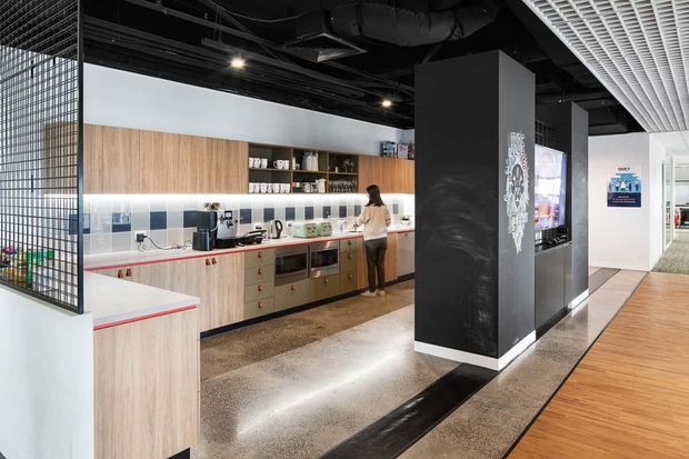 A contemporary commercial interior featuring an open kitchenette or staff break area with light timber cabinetry, white benchtops, tiled backsplash, integrated appliances and open shelving. Black exposed services and ceiling elements contrast with a white linear ceiling grid. A polished concrete floor and timber-finished circulation area lead past dark feature columns and display screens.