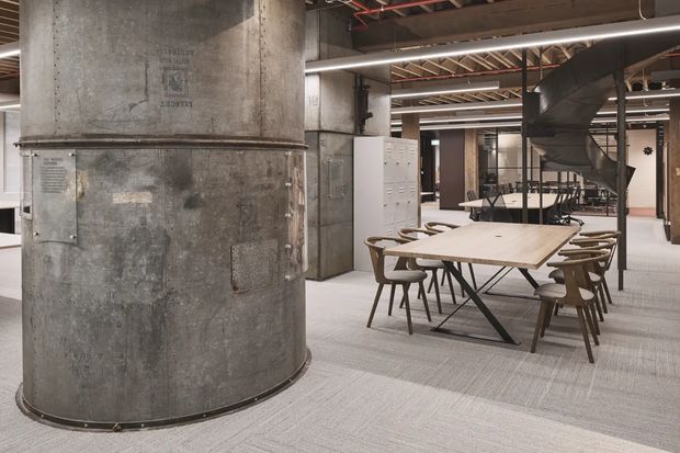 An open-plan commercial interior featuring a large meeting or communal dining table surrounded by timber-framed chairs. The space has exposed industrial services, including large metal ventilation ducts, visible ceiling structure, concrete-look cylindrical columns, metal-framed glazing, and light grey carpet. The overall appearance is a refined industrial office or hospitality workplace.