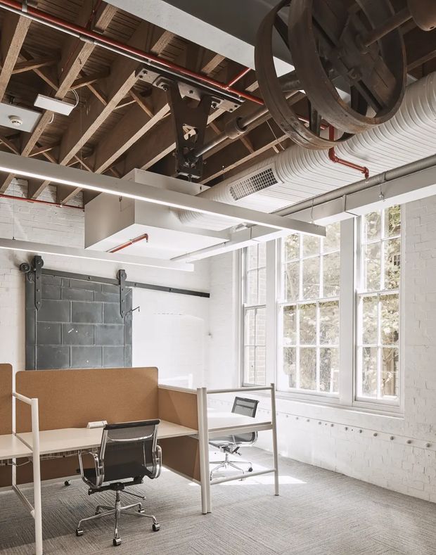Bright open-plan office interior with shared workstation desks, tan acoustic privacy screens, rolling office chairs, and a large industrial-style sliding barn door. White-painted brick walls, tall multi-pane windows, exposed timber roof trusses, visible ductwork, and exposed red sprinkler piping create a converted warehouse or loft-office character.