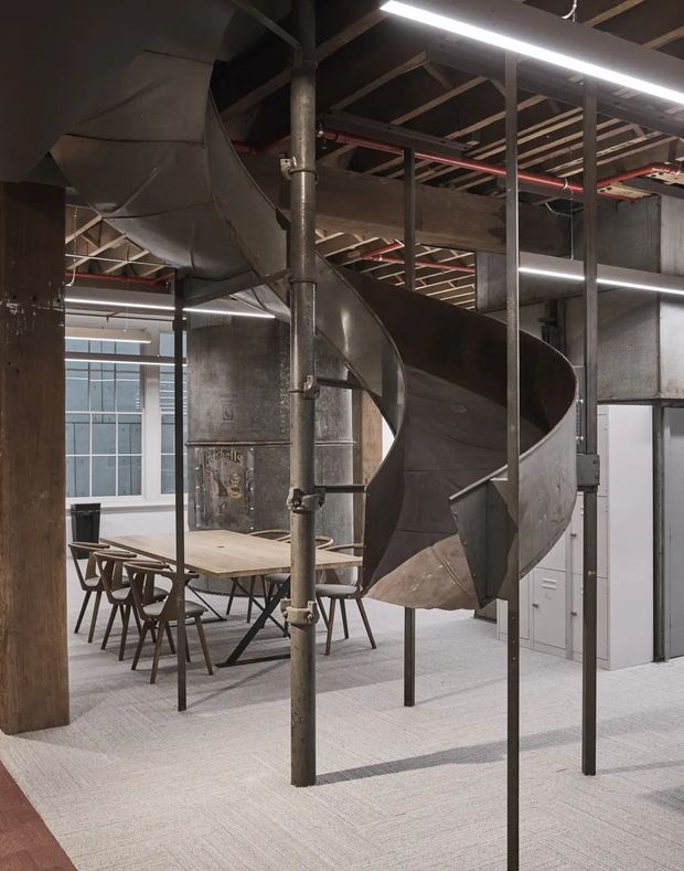 An industrial-style commercial interior featuring a large sculptural metal spiral staircase or slide-like stair structure supported by slender steel posts. The space has exposed roof framing, visible mechanical ductwork and pipes, concrete and dark metal surfaces, linear fluorescent lighting, grey carpet, and a timber communal dining or meeting table with wooden chairs. Filing cabinets and glazed partitioning are visible in the background.