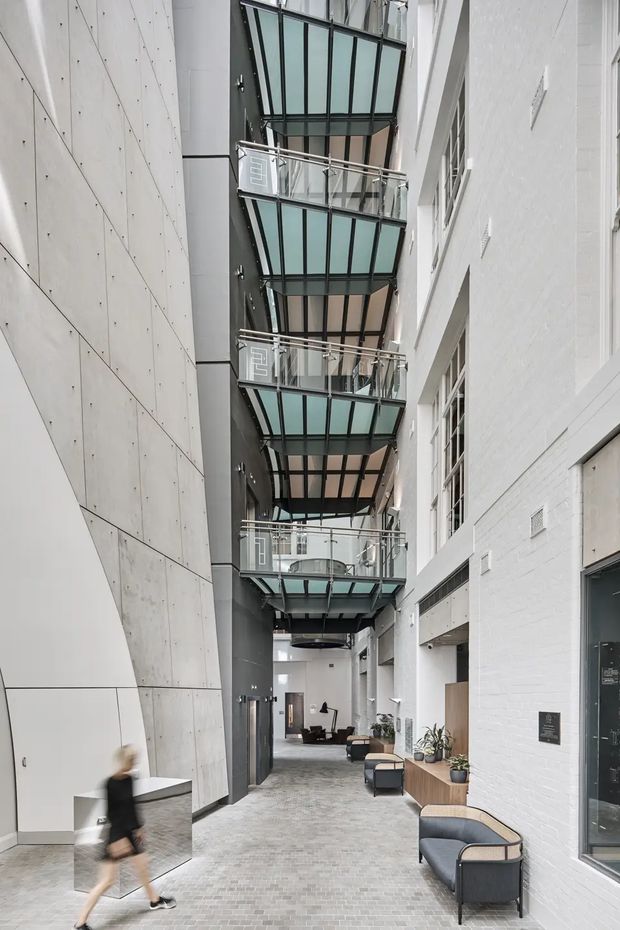 A tall, narrow contemporary commercial atrium or internal laneway with exposed concrete and white-painted brick walls, a grey paver floor, and multiple projecting glass-and-metal balconies stacked beneath a glazed roof structure. Lounge seating, planters, doors and windows line the ground level, creating a bright modern communal circulation space.