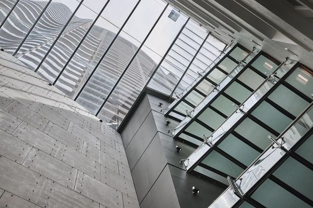 Upward-looking view within a large contemporary atrium, showing exposed smooth concrete walls, extensive glass roof glazing, dark metal-framed internal glazing or balconies, and a sculptural rippled facade visible beyond the roof. The composition is strongly vertical and geometric, with a cool grey architectural palette and abundant daylight.