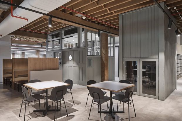 A contemporary commercial interior with shared dining or breakout tables, booth seating, and a compact enclosed meeting or office pod. The space features exposed timber roof framing, visible services and red fire-suppression pipes, polished concrete flooring, dark grey vertically panelled walls, extensive internal glazing, and warm timber partitions. The overall aesthetic is industrial and utilitarian with an open-plan workplace character.