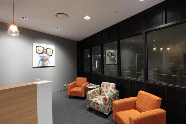 A compact, contemporary reception or waiting area with orange upholstered armchairs and a colourful patterned lounge chair arranged around a small table. A white reception desk with timber frontage sits at left, while dark full-height glazed joinery forms a partition along the rear wall. The space has dark grey walls, carpet flooring, recessed downlights, and a suspended pendant light, creating a refined but welcoming commercial interior.