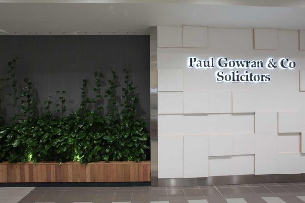 A commercial lobby or entry area featuring an illuminated “Paul Gowran & Co Solicitors” sign mounted on a white geometric wall, beside a built-in planter with dense greenery. The space has a contemporary, polished appearance with dark vertical wall panelling, timber planter cladding, and tiled flooring.
