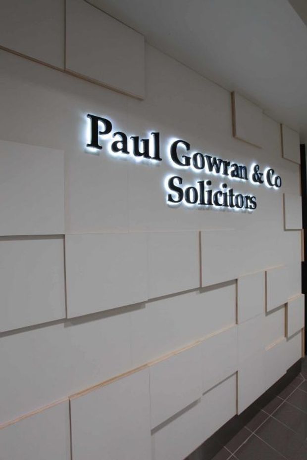A contemporary commercial law firm reception or entry wall featuring an illuminated black sign reading “Paul Gowran & Co Solicitors”. The wall is clad in a geometric arrangement of white rectangular panels with narrow shadow gaps and subtle timber-toned edging. A dark tiled floor and dark skirting provide contrast. The image is tightly framed on the branded feature wall, with no furniture visible.