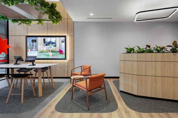 Contemporary commercial reception or waiting area with a small meeting table, lounge chairs, timber-look wall panelling, a large built-in planter cabinet, geometric grey feature wall, integrated display screen, and suspended greenery. The space has warm timber tones, soft grey carpet zones, and refined modern lighting.