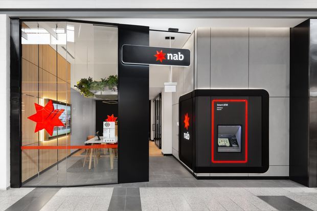 A contemporary bank branch entrance viewed from a shopping-centre or commercial corridor. The frontage features full-height glass doors and glazing, black framing and wall panels, grey tiled flooring, and prominent red-and-black NAB branding. An ATM is integrated into a rounded black wall enclosure on the right, while the interior shows a reception or consultation area with timber wall cladding, greenery, and a corridor beyond.