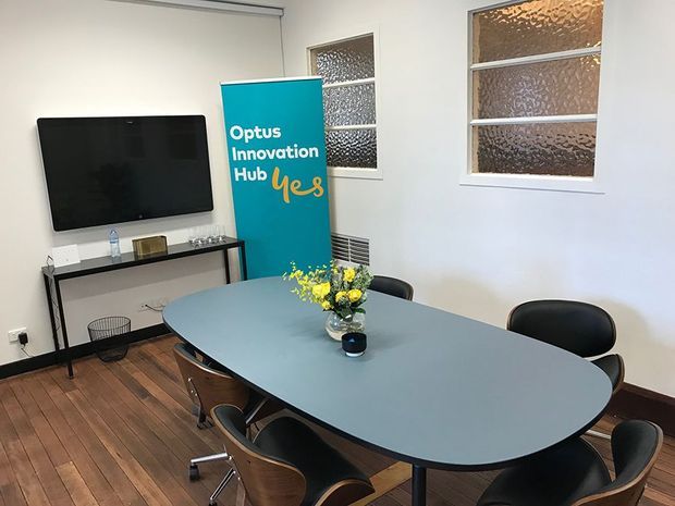 A compact meeting room with a large oval conference table surrounded by office chairs, a wall-mounted display, a slim console table, and an Optus Innovation Hub banner. The room has white walls, timber flooring, textured glass windows, and simple functional office furnishings.