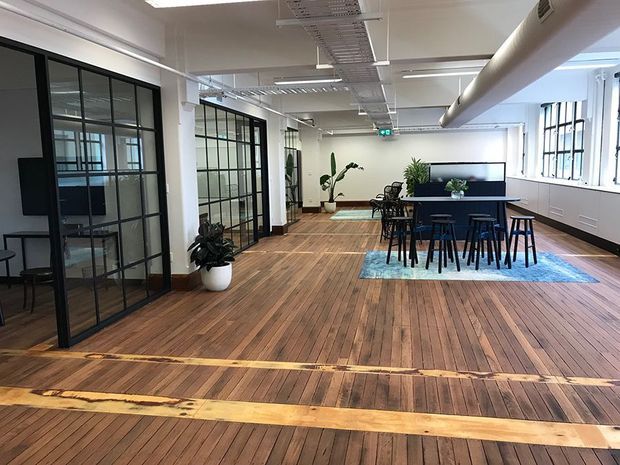 Open-plan commercial interior with exposed timber floorboards, black-framed glazed meeting rooms, white structural columns, exposed services, and a communal dining or breakout area with tables, stools, a large screen, and indoor plants. The space has an industrial office or warehouse-conversion character with abundant daylight.