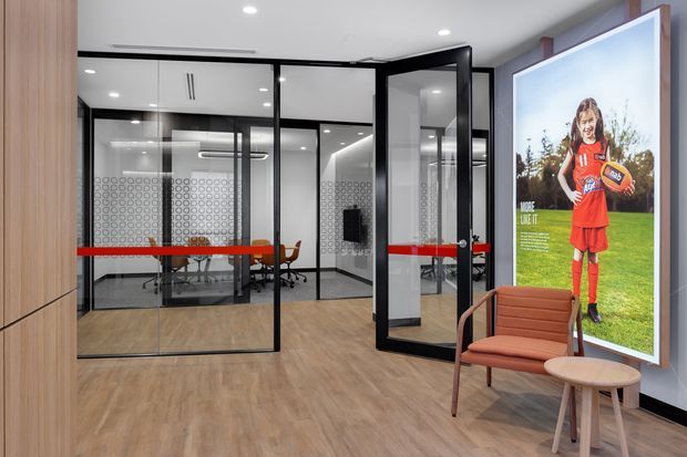 Contemporary commercial office reception or waiting area with black-framed glass-partitioned meeting rooms, warm timber-look flooring and wall panelling, recessed ceiling lighting, and a large sports-themed graphic feature panel. A rust-coloured lounge chair and small round side table create a compact seating zone in the foreground.