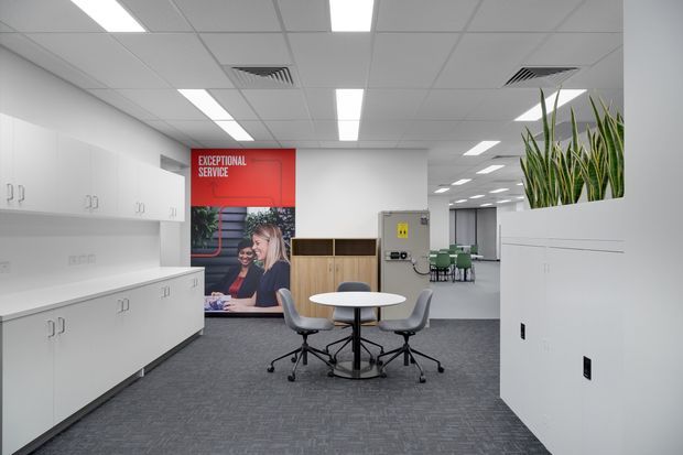 A bright contemporary commercial office interior with white built-in cabinetry and work surfaces, grey carpet tiles, a suspended acoustic tile ceiling with recessed linear lighting, and a small round meeting table with rolling chairs. A reception or collaboration area is visible beyond, with a branded wall graphic and indoor planting integrated into the cabinetry.
