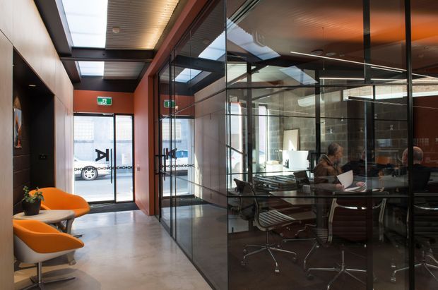 A contemporary commercial office interior showing a glazed meeting room with a long conference table and office chairs, viewed from a circulation corridor. The corridor features orange lounge chairs, a small round table, dark built-in wall panels, polished concrete flooring, timber-look ceiling lining, exposed services, and skylights. Full-height black-framed glazing creates strong visual separation while maintaining transparency between the corridor and meeting space.