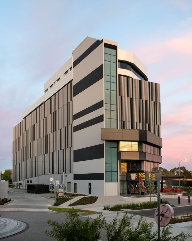 Exterior architectural photograph of a contemporary multi-storey commercial building at dusk. The building has a long rectilinear form, a prominent glazed vertical stair or circulation tower, alternating dark and light horizontal bands, and extensive vertical fins screening the facade. A retail or commercial tenancy occupies the lower frontage, with landscaped paths, street furniture, signage, and a street setting in the foreground.
