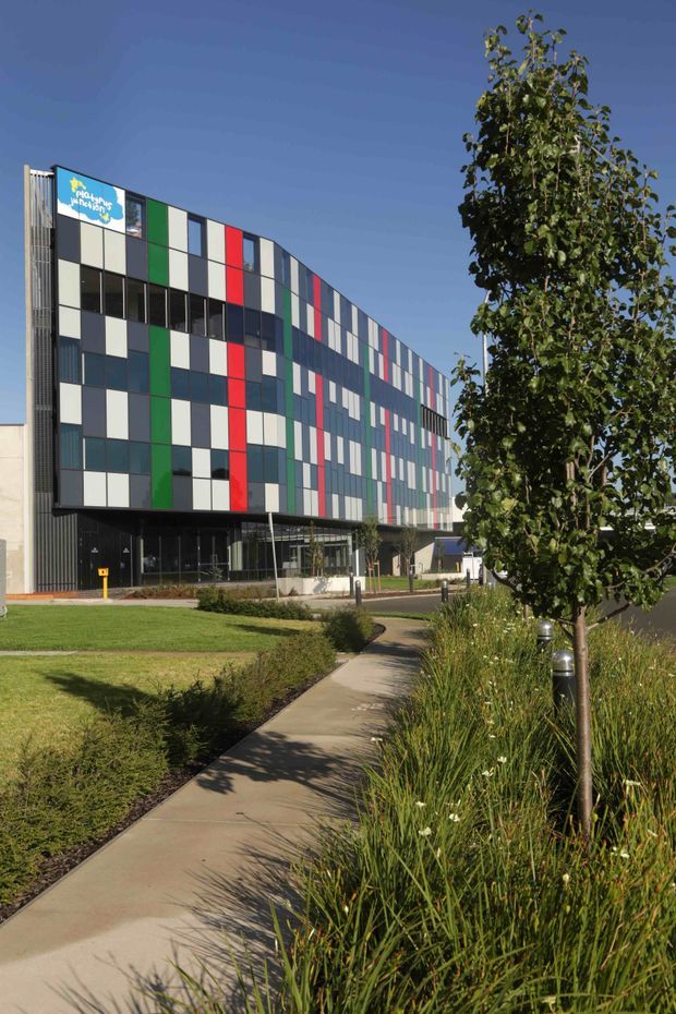 Exterior view of a contemporary multi-storey commercial building with a curved facade composed of alternating dark glazing and colourful rectangular panels in white, grey, green, red and blue. The building sits within landscaped grounds with a paved pedestrian walkway, lawn and ornamental grasses, under a clear blue sky.