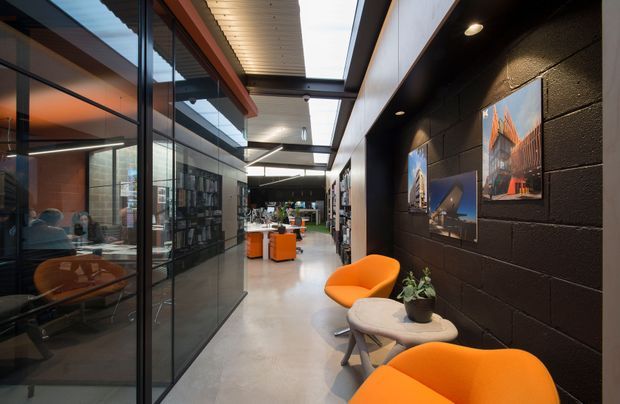 A contemporary commercial interior showing a polished concrete corridor or lounge beside glass-partitioned offices and meeting rooms. The space features a black painted block feature wall displaying architectural photographs, exposed structural and service elements, linear skylight-like glazing, recessed lighting, and vivid orange lounge chairs with a small round table and plant. Workspaces and additional seating are visible deeper into the office.