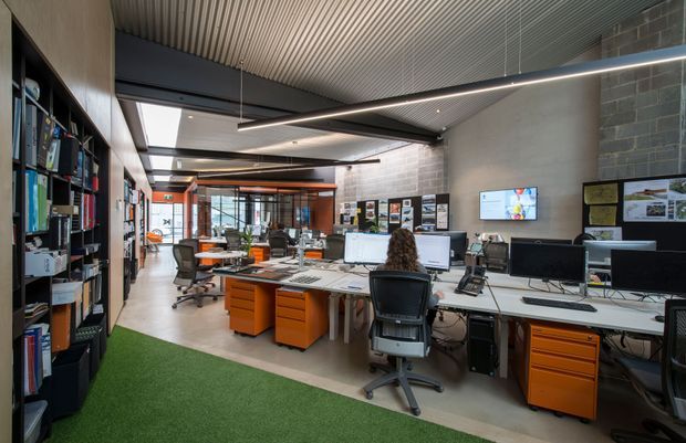 Open-plan contemporary office interior with multiple workstations, computers, black ergonomic office chairs, orange mobile drawer units, shelving filled with books and files, display boards, and linear suspended lighting. The space has a ribbed metal ceiling, polished concrete flooring, dark masonry feature walls, extensive glazing at the far end, and a strip of green carpet or artificial turf in the foreground.