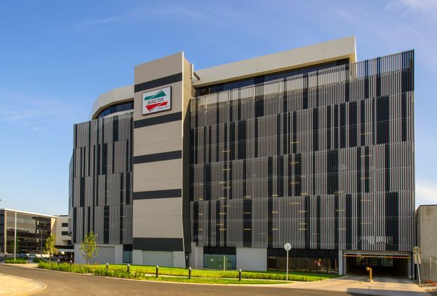 Exterior view of a contemporary multi-storey commercial building with a prominent vertical metal screening façade, dark glazed window bands, and a tall beige and charcoal sign tower. Landscaped frontage and a covered vehicle entry are visible under a clear blue sky.