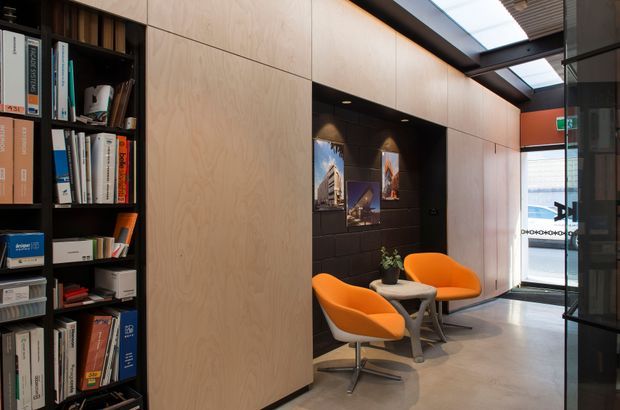 A contemporary commercial reception or waiting area with built-in open shelving filled with books and files, large pale timber wall panelling, a dark tiled feature alcove displaying framed artwork, and two orange lounge chairs around a small round table. The space has a polished concrete floor, exposed dark ceiling structure with skylights, and glazed entry doors at the far end.