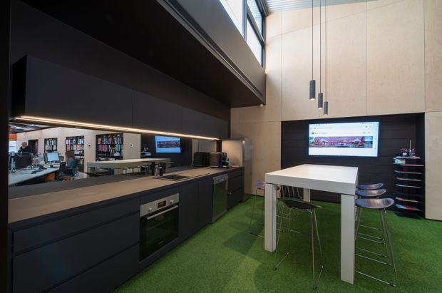 A contemporary open-plan kitchen and dining workspace with dark matte cabinetry, integrated appliances, a long reflective splashback, and a white island or dining table with bar stools. The room has a dramatic double-height volume, pale timber-toned wall panels, a dark feature wall, suspended pendant lights, high clerestory glazing, and vivid green carpet flooring. A reflected office or library workspace is visible in the splashback.