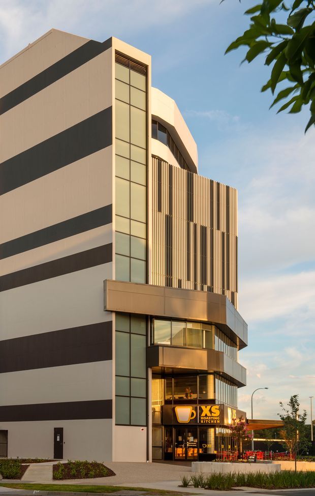 Exterior architectural photograph of a contemporary multi-storey commercial building. The facade combines broad alternating dark and light horizontal bands, a tall glazed vertical stair or circulation element, curved projecting balconies, and a prominent vertical timber-toned screening system. A ground-floor XS Roasting Kitchen restaurant tenancy opens onto a landscaped forecourt and outdoor seating area.