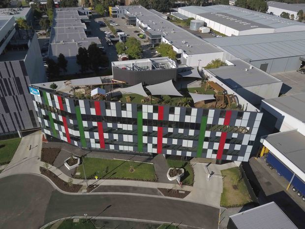 Aerial view of a contemporary multi-storey commercial or institutional building in an industrial precinct. The curved facade is clad in a bold geometric grid of dark grey, white, red, and green panels, with extensive glazing. Landscaped grounds, paved access paths, rooftop terraces with shade sails, and surrounding large industrial buildings are visible.