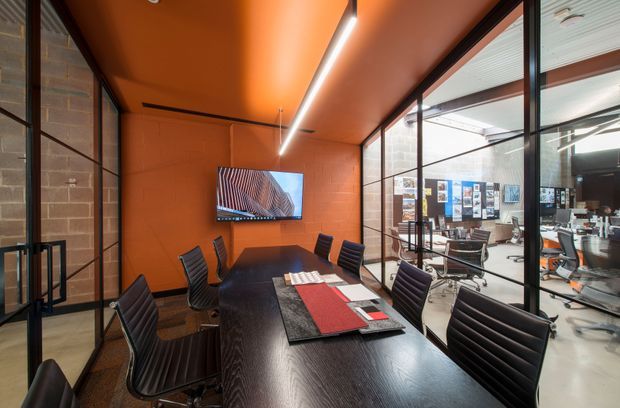 A contemporary meeting room with a long dark wood conference table and black ribbed office chairs. The room features vivid orange painted walls and ceiling, integrated linear lighting, exposed brick accents, and extensive black-framed internal glazing overlooking an open-plan office area. A wall-mounted display and presentation materials reinforce the commercial workplace setting.