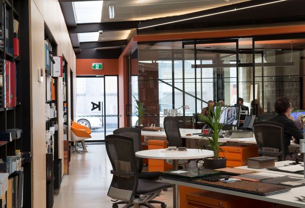 A contemporary commercial office interior with open-plan workstations, black mesh office chairs, orange built-in desks and storage, glass-partitioned meeting or office rooms, and a small round collaboration table. The space features exposed dark ceiling services, linear lighting, timber-toned wall panels, shelving, and abundant glazed partitions, creating an industrial-modern workplace atmosphere.