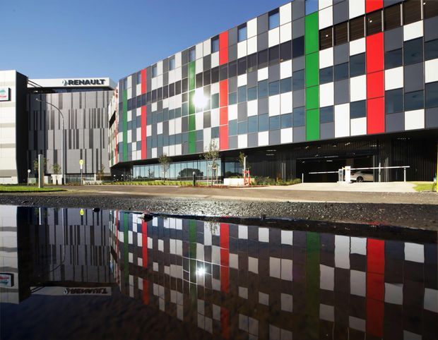 Exterior view of a contemporary multi-storey commercial building with a highly patterned facade of white, grey, black, red, green and blue rectangular panels. A Renault-branded building is visible to the left, with glazed ground-level frontage, a covered entry or parking area, landscaping and strong reflections in a large foreground puddle.