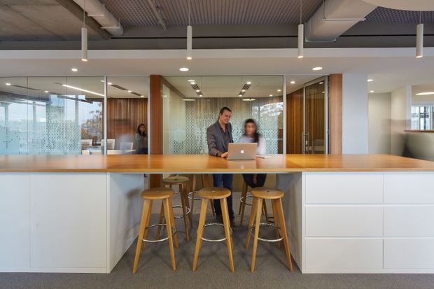 A contemporary commercial office breakout and collaboration area featuring a long timber-topped island bench with integrated white cabinetry and four timber bar stools. Two people work at a laptop behind the bench. Glazed meeting rooms with patterned privacy film, timber wall panelling, exposed services, suspended linear lighting, and a carpeted floor create a modern workplace setting.