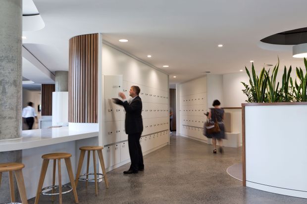 Bright contemporary commercial lobby or reception area with extensive white built-in locker or storage cabinetry, a polished concrete floor, cylindrical concrete columns, and curved white counters. Vertical timber slats form a feature partition, with timber stools at a counter and integrated planter boxes containing tall greenery. People are present, suggesting an active workplace or public facility.