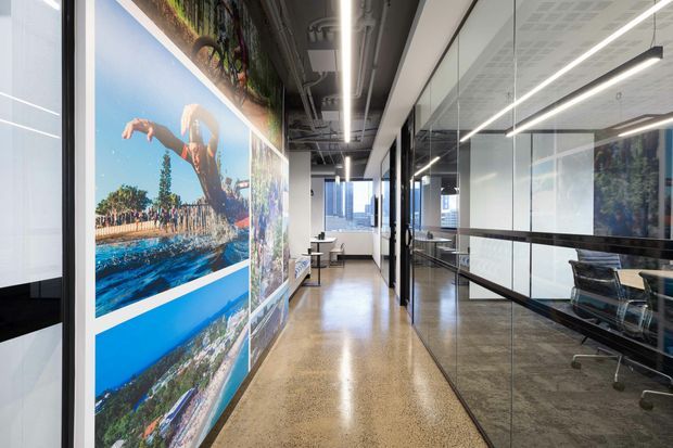 A bright contemporary commercial office corridor with polished concrete flooring, glass-partitioned meeting rooms, linear suspended lighting, exposed services, and large photographic wall graphics depicting coastal scenes. Workstations and office furniture are visible through the glazed partitions.