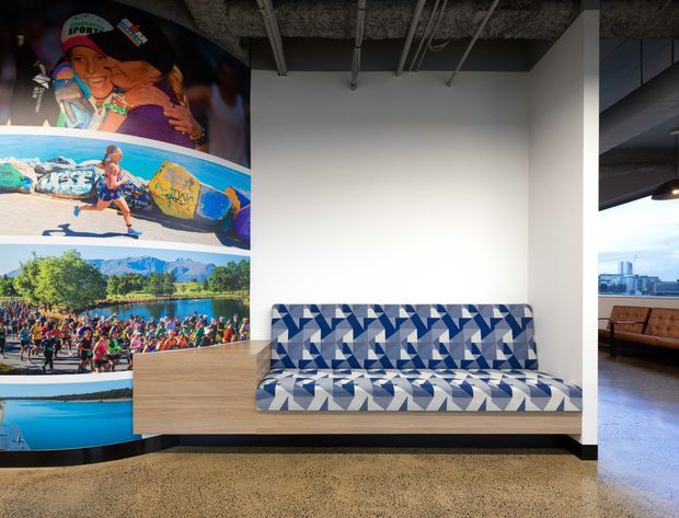 A contemporary commercial lobby or waiting area featuring a bold geometric blue, white, and grey upholstered bench against a plain white wall. A timber-clad built-in counter or cabinet sits beside the seating, while a large colourful photographic mural wraps around the left wall. The exposed dark ceiling with visible services and polished concrete floor create an industrial-style fit-out. An adjoining lounge area and city view are visible through the opening on the right.