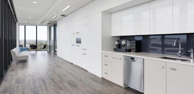 A bright, open-plan contemporary kitchen with extensive glossy white handleless cabinetry, a dark reflective splashback, pale stone-look benchtops, and stainless-steel appliances. The kitchen opens into a spacious living area with grey timber-look flooring, floor-to-ceiling glazing, and a black vertical feature wall.