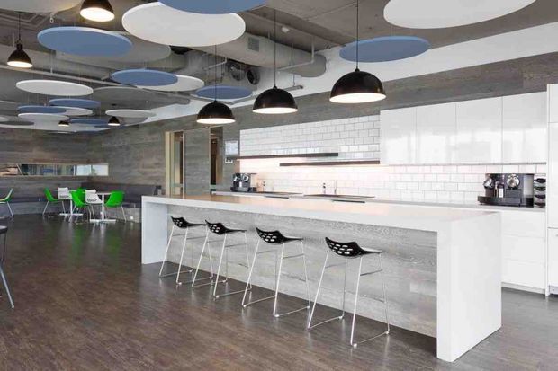 A spacious contemporary commercial-style kitchen and dining area with a long white island, integrated seating, black bar stools, dark timber flooring, white cabinetry, and a white brick-tile splashback. Exposed concrete ceiling services and ductwork are paired with suspended black pendant lights and layered blue, white, and grey acoustic ceiling panels. A small casual dining or breakout area is visible in the background.