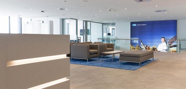A spacious contemporary commercial interior, likely a reception or lounge area, with a large illuminated partition or reception desk in the foreground. The space features neutral carpeted flooring, grey upholstered lounge seating arranged on a blue rug, glass balustrades and partitions, extensive glazing, and a large blue digital display wall.