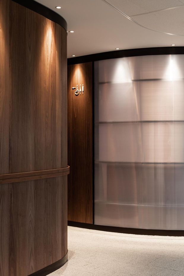 A refined commercial interior lobby or reception area featuring a curved dark timber-paneled wall in the foreground, dark wood wall panels with illuminated signage, a large translucent ribbed or textured partition, recessed ceiling lighting, and pale carpet flooring. The palette is warm wood, black trim, soft metallic beige, and cream, creating a polished hospitality or corporate atmosphere.
