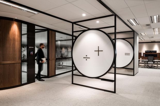 A contemporary commercial office interior with a spacious reception or circulation area. Black-framed glazed partitions create meeting rooms and feature oversized circular white panels marked with minimalist mathematical symbols. Curved dark timber wall panelling and doors contrast with the light carpet, white suspended ceiling, and exposed linear office lighting. Workstations and office furniture are visible in the background.