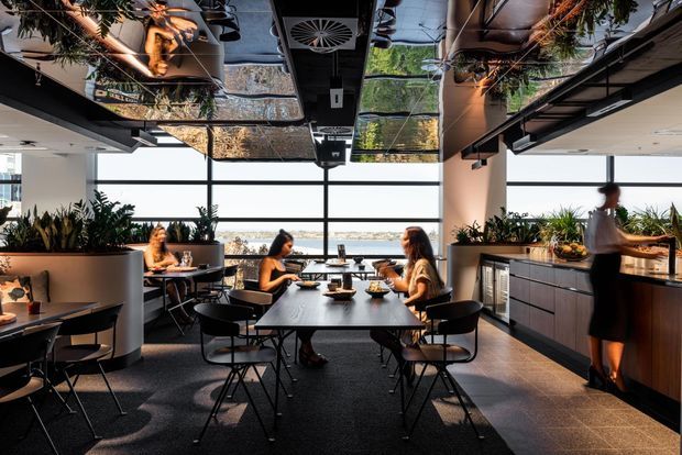 A contemporary dining area in a bright commercial hospitality space, with multiple tables and black chairs arranged beside full-height windows overlooking water and a distant landscape. A service counter and fixed cabinetry line the right side, while large planters and abundant greenery divide seating zones. The ceiling features reflective panels, exposed services, suspended lighting, and hanging plants, creating a modern, polished restaurant or café interior.