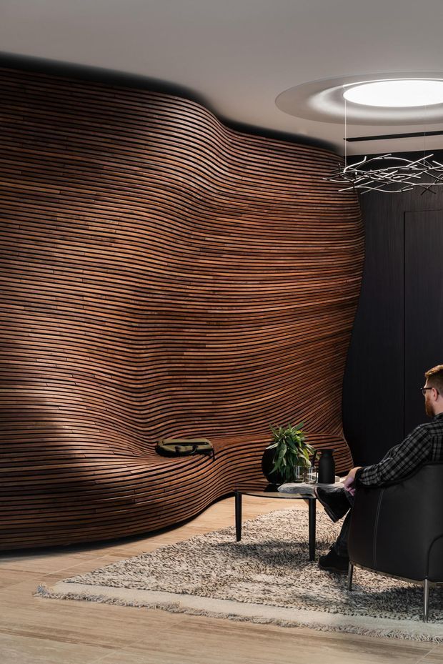 A sophisticated contemporary interior lounge or reception space featuring a dramatic organically curved wall clad in continuous horizontal dark timber slats. The room includes a black lounge chair, low round coffee table, textured area rug, potted plant, dark vertical joinery, and a sculptural suspended light beneath a recessed circular ceiling feature. The palette is warm timber, black, grey, and muted neutral tones, creating a refined, moody atmosphere.
