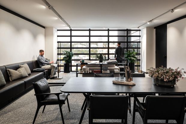A contemporary open-plan office lounge and meeting area with a large black-framed glazed wall, linear track lighting, black leather seating, a long dark meeting table, and indoor plants. Two people occupy the space, with a low media or display console positioned in front of the windows. The palette is monochromatic and refined, with concrete-like grey flooring, white walls, and black architectural detailing.