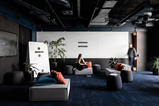 A dark, contemporary shared lounge or breakout area with modular sofas, ottomans, cushions, indoor plants, and a large wall-mounted screen. The room has a dramatic exposed-services ceiling with black ductwork, suspended lighting, and visible mechanical systems. White and charcoal wall panelling, deep blue carpet, black furniture, and blue structural columns create a moody, refined commercial interior.